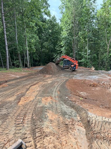 A red excavator moves dirt on a construction site surrounded by trees, with visible tire tracks on the ground.