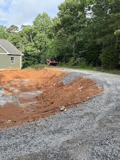 A gravel driveway curves around a mound of red dirt near a partially visible house, with dense green trees in the background. A vehicle is parked along the driveway.